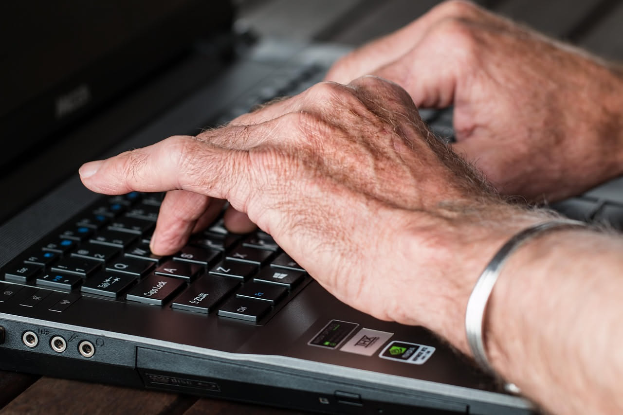 Anziano seduto al tavolo con tablet acceso, mani poggia sullo schermo, occhiali da lettura, ambiente domestico caldo con finestra sullo sfondo
