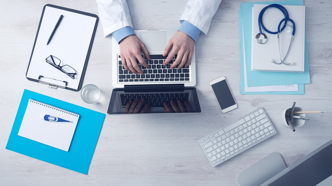 Persona che scrive una mail su un laptop con in primo piano uno schermo che mostra un messaggio email aperto, con accanto un termometro e appunti medici sfocati sullo sfondo