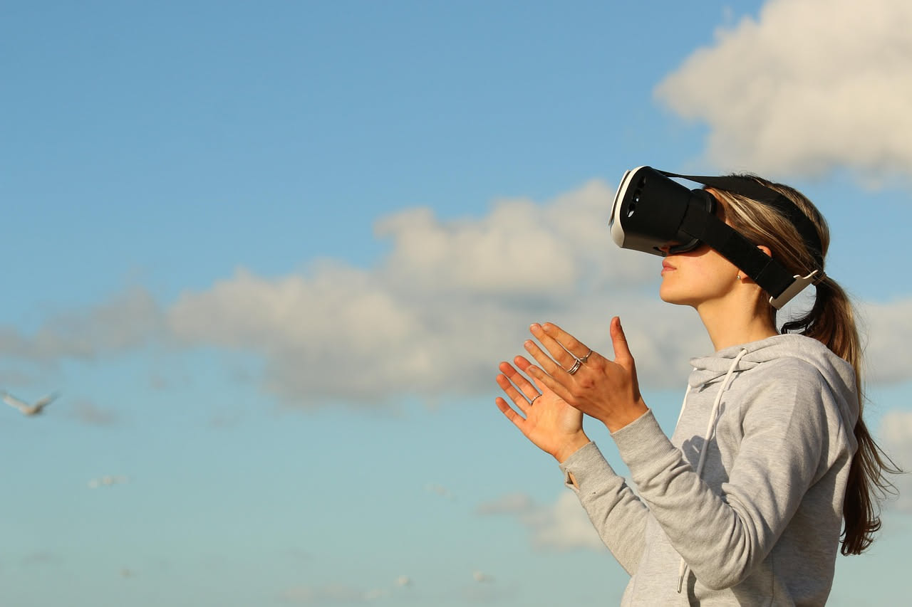 Persona che indossa un visore di realtà virtuale nero con due controller bianchi nelle mani, immersa in uno spazio luminoso con ambiente virtuale sullo sfondo sfocato