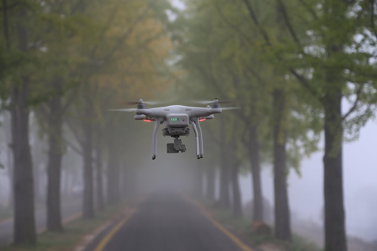 Drone quadricottero in volo con quattro eliche visibili, sfondo cielo azzurro, vista dall'angolo inferiore che mostra il sottocarro e i sensori